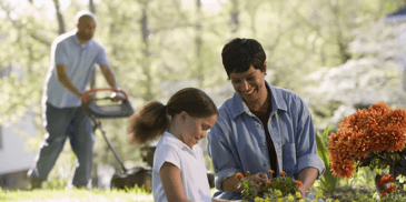 a woman and a child smiling in front while a man is mowing the lawn in the background